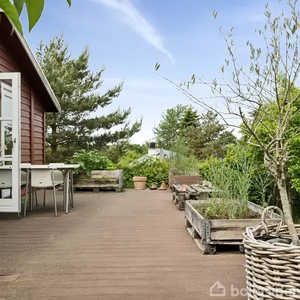 Et træhus med hvid dør åbner ud til en træterrasse. Havemøbler står ved siden af plantekasser og grønne omgivelser, omkranset af træer og buske under en klar blå himmel.