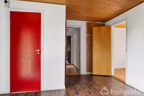 Rød dør står lukket mod en hvid væg, mens en trædør står åben. Gulv belagt med brune fliser, og loft er af lyst træ. Boligsiden-branding ignoreret.