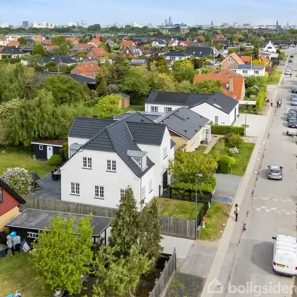 Hvidt hus med sort tag står blandt andre villaer. En bil kører på en gade omgivet af grønne haver og træer i forstæderne, en klar himmel ses i baggrunden.
