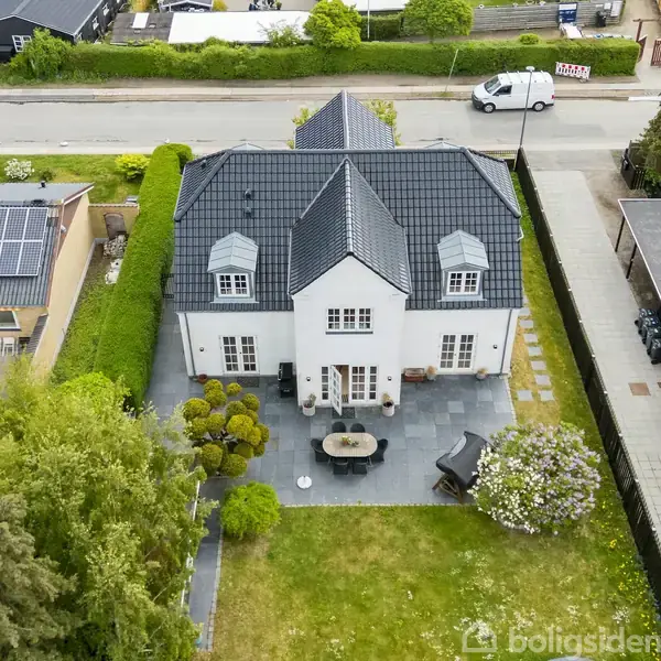 Hvidt hus med sort tag ses fra oven, omgivet af grøn have og nabobygninger. Foran huset er der en terrasse med bord og stole. En vej løber foran ejendommen.