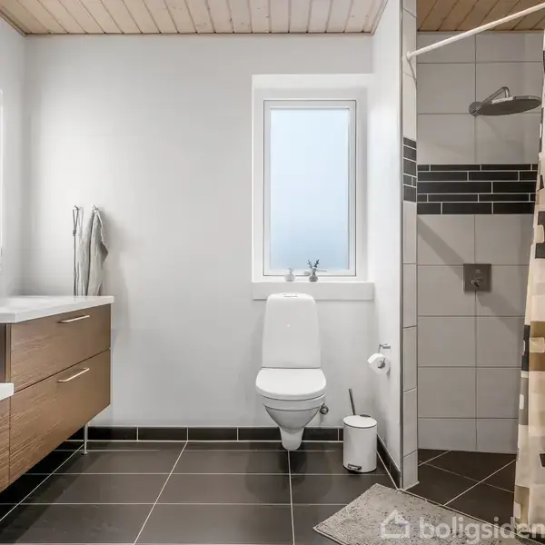 Toilet står ved siden af et hvidt vindue, omgivet af et moderne badeværelse med bruseområde til højre og dobbeltvask med spejl til venstre. Mørke fliser dækker gulvet.
