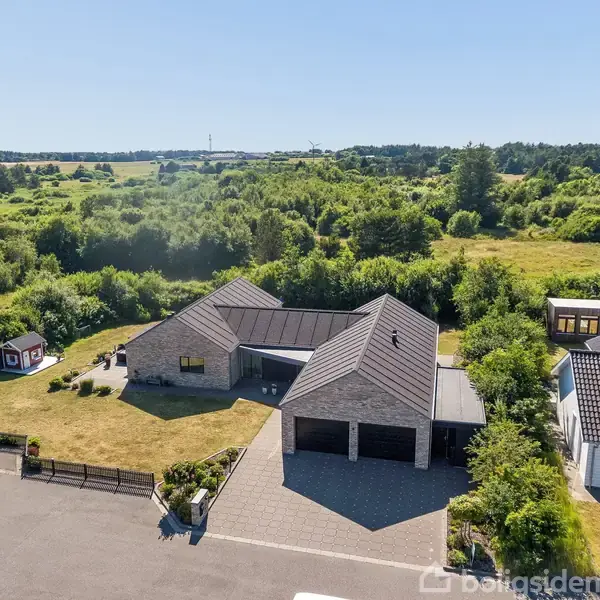 Et moderne hus med en grå tagkonstruktion og tilhørende have står stille på en stille boligvej. Det er omgivet af frodigt, grønt landskab og nabohuse under en klar blå himmel.