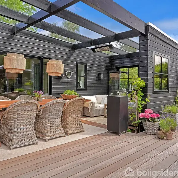 Terrasse med træbord og flettede stole står under en åben pergola. Omkring er der planter i krukker, og terrassen er tilstødende et moderne, sort træhus med store vinduer.