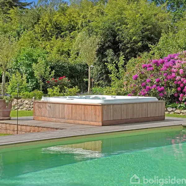 Boblebad står stille ved siden af en swimmingpool omgivet af frodig grønne buske og blomstrende planter. Den fredfyldte have skaber en afslappet atmosfære.