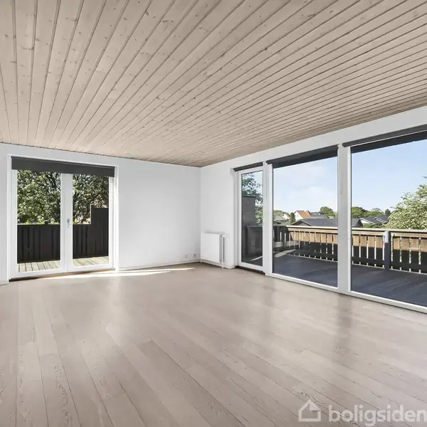 Rummeligt rum med trægulv og lyse vægge har store vinduer og dør ud til en balkon med udsigt over et grønt område med træer.