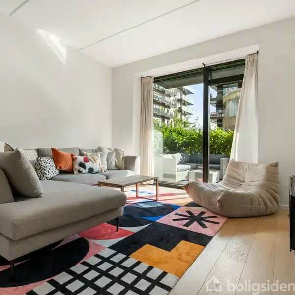 En grå hjørnesofa med farverige puder står i en lys stue. Sofaen vender mod en tv-skærm, og en glasskydedør åbner ud til en terrasse med grønne planter.