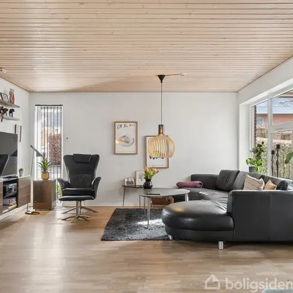 Sort lædersofa står i stue, omkranset af store vinduer. Et sofabord med lampe hænger over. Der er også en lænestol, et fjernsyn og flere små dekorationer.