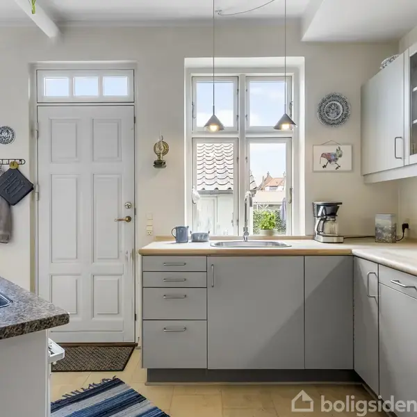 Et lyst køkken med hvide skabe og grå skuffefronter. Kaffen står klar ved kaffemaskinen på bordpladen. Vinduerne viser tegltage udenfor. Blå stribede tæpper på gulvet.