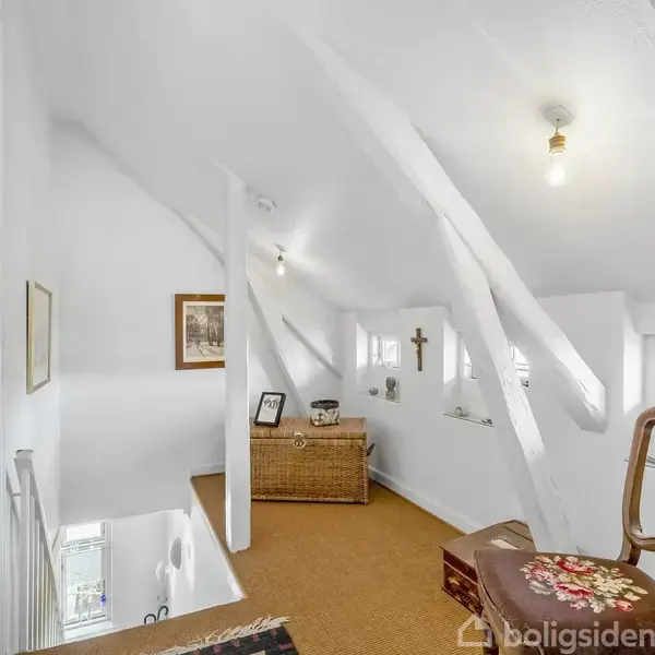 En lys trappeopgang med synlige træbjælker og hvidmalede vægge. En stol og en kurvekiste står i rummet, der er dekoreret med billeder og kors på væggene.