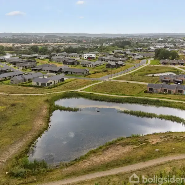 En lille sø omgivet af grønne græsarealer. I baggrunden ses en række tætliggende huse og veje i en beboelsesudvikling. Landskabet er fladt med spredt vegetation.