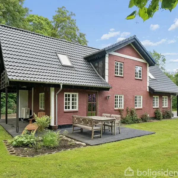 Et rødt hus med sort tag står omgivet af grønt græs og træer. Foran huset er der en terrasse med bord og stole.