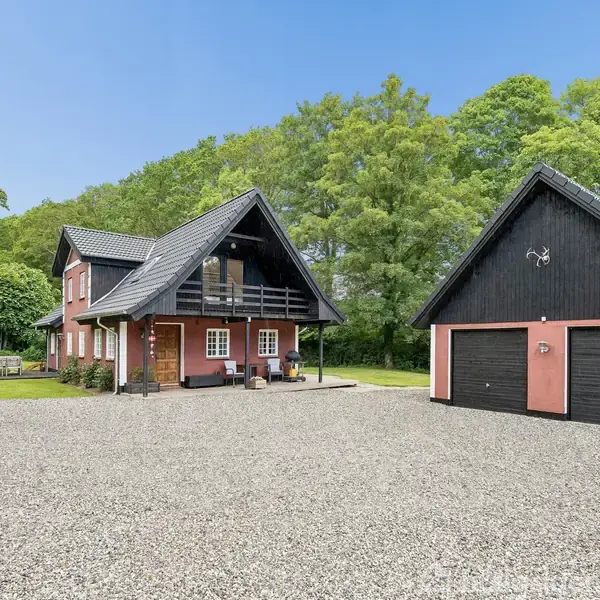 Et rødt landhus med sort tag står på en grussti. Ved siden af er en garage i samme stil. Omgivet af grønne træer og græsarealer.