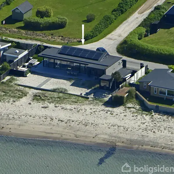 Hus med solpaneler på taget står stille ved kysten omgivet af andre huse og grønne marker, mens en vej løber bag husene.