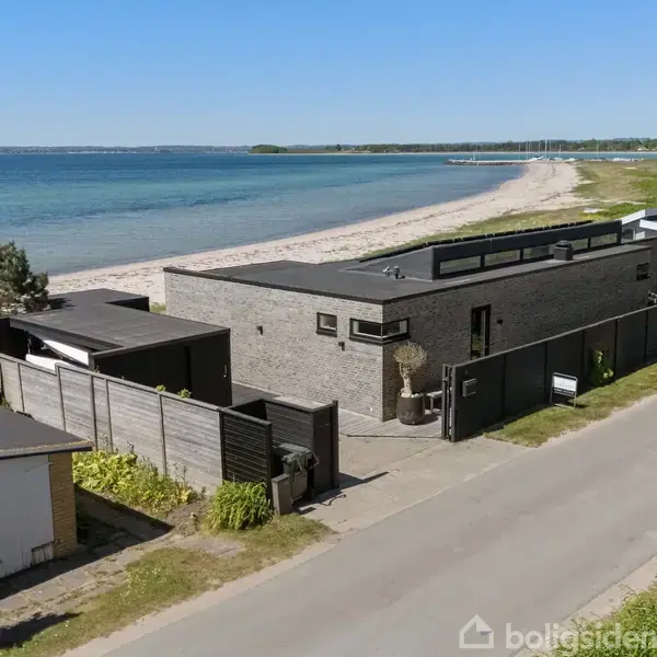 Et moderne, minimalistisk hus ligger langs en bred vej ved kysten. Huset har fladt tag og grå murstensvægge. Kystlinjen og havet ses i baggrunden under en klar blå himmel.