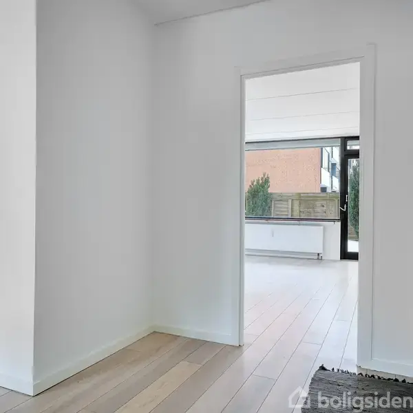 En åben dør fører ind til et lysfyldt rum med trælaminatgulve. I baggrunden ses store vinduer og en glasdør med udsigt til en terrasse og træhegn.