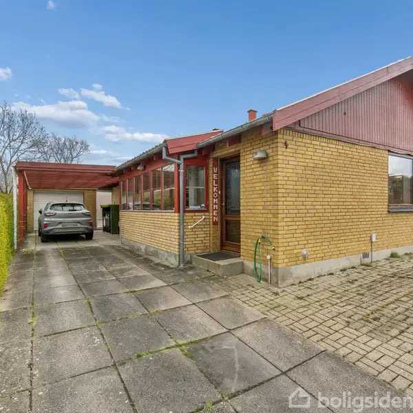 En gul murstensvilla med rødt tag står ved en flisebelagt indkørsel. En bil er parkeret under en carport. Hækken ved siden af indkørslen er frodig og grøn.