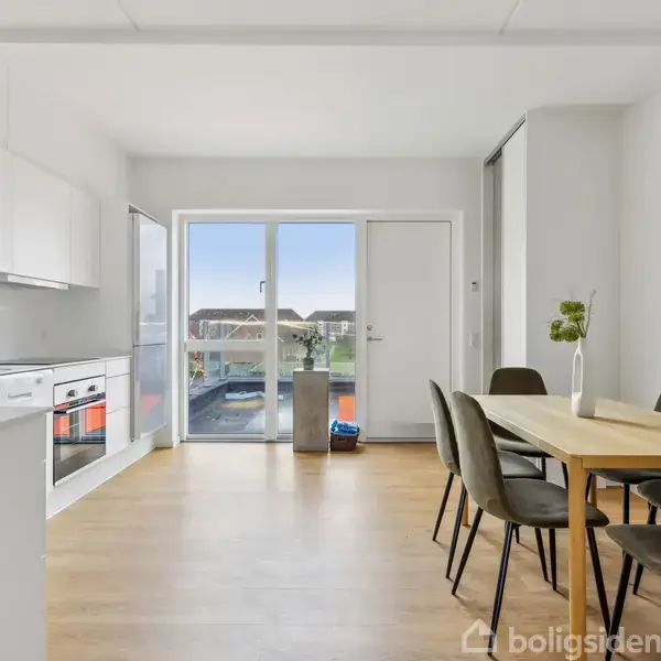 Et lyst køkken-alrum med trægulv indeholder et spisebord og seks stole. Køkkenet har hvide skabe og en ovn. Store glasdøre åbner ud til en balkon med udsigt.