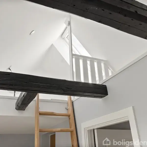 Et lyst værelse med trægulv og en åben dør til en balkon. En stige fører opad, og et lille bord med lampe står i hjørnet.