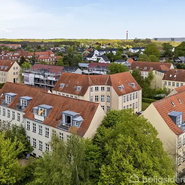 Åbent glasdør fører ud til en balkon med udsigt over et boligområde med røde tage og grønne trækroner. Indendørs ses et lyst trægulv og en radiator.