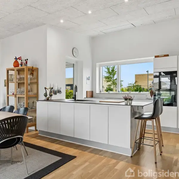 Køkken i moderne stil med hvidt skab og køkkenø. Høj stol ved køkkenøen. Spisebord med stole, hængelamper over bordet. Lys fra stort vindue. Trægulv.