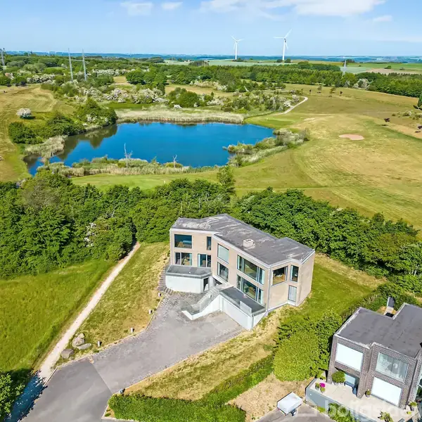 Moderne hus står stille mellem grønne marker og træer. I baggrunden ses en sø omkranset af natur og vindmøller under en blå himmel.