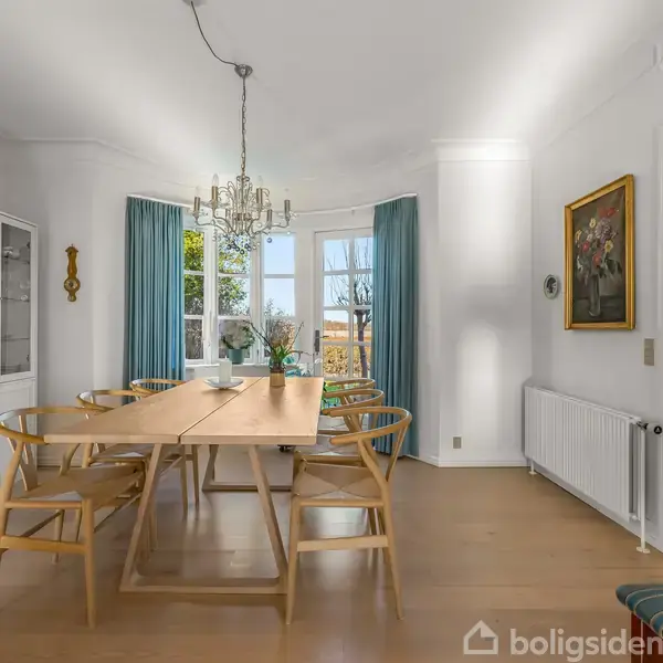 Et spisebord med seks stole står oplyst af en lysekrone. Store vinduer med blå gardiner viser en have. Rummet har hvide vægge, indbygget bogreol og træmøbler.