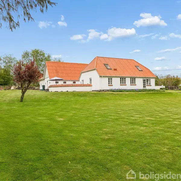Et hvidt hus med rødt tag står midt på en grøn græsplæne omgivet af træer og buske under en blå himmel med skyer.