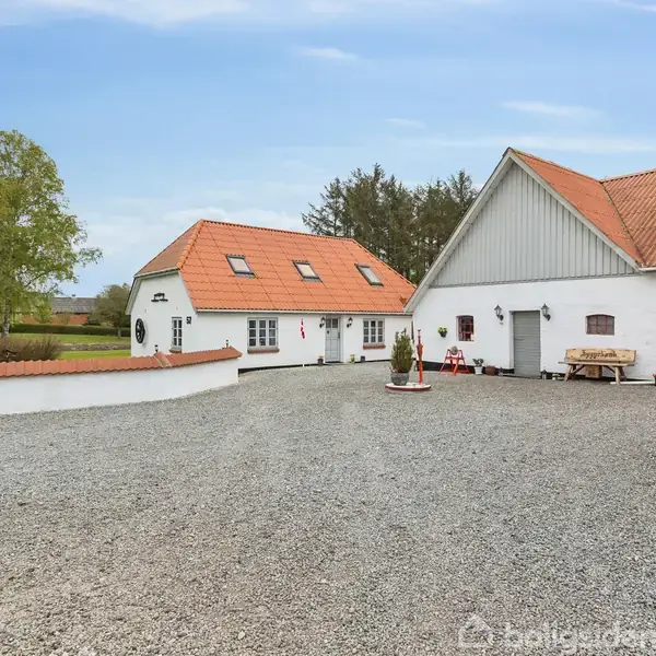 Et hvidt hus med rødt tegltag står stille på en grusbelagt gårdsplads. Der er grønne træer og en lav mur omkring. Der ses en lade med åben port.