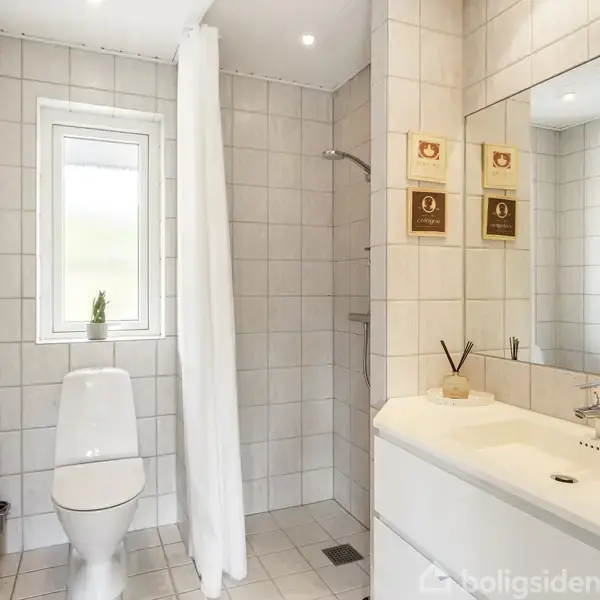 Toilet er placeret ved siden af et lille vindue, med en bruser til højre skjult af et hvidt forhæng. En hvid håndvask ligger på en bred bordplade til højre.