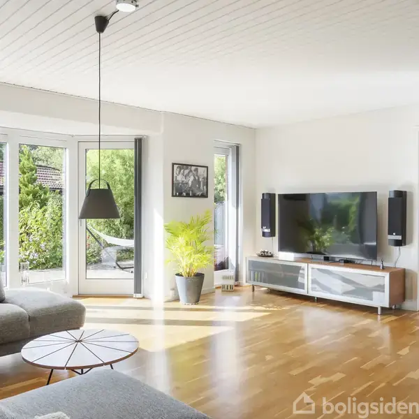 Stue med grå sofa, rundt sofabord og fladskærms-TV på hvid skænk. Store vinduer giver lys og udsigt til haven. Skandinavisk indretning med en grøn plante ved siden af TV'et.