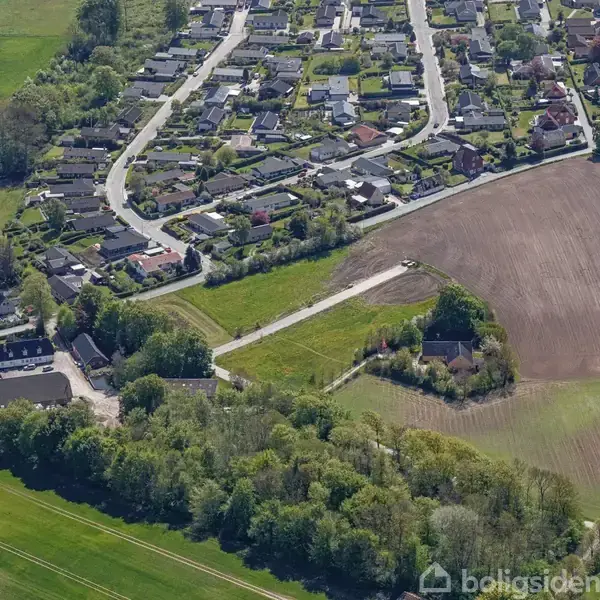 Boligområde med mange huse ligger ved siden af et åbent markområde. Veje snor sig gennem boligområdet, og grønne træer omgiver stedet, hvilket skaber en kontrast mellem bebyggelse og natur.