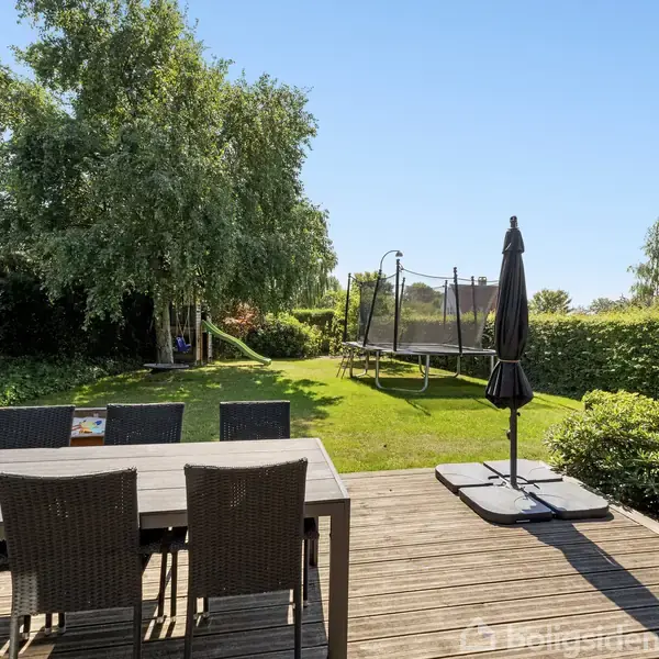 Terrasse med et bord og stole står på et trædæk. En stor, sort parasol er foldet sammen. I baggrunden er en grøn græsplæne, trampolin og buske. En træ fylder venstre hjørne.
