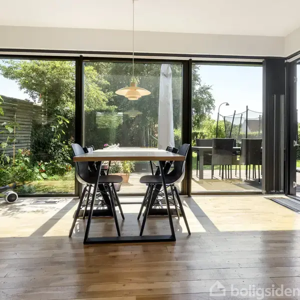 Et spisebord med stole står foran store glasdøre, der åbner ud til en terrasse og grøn have. Sollys stråler ind, og en brændeovn ses til højre.