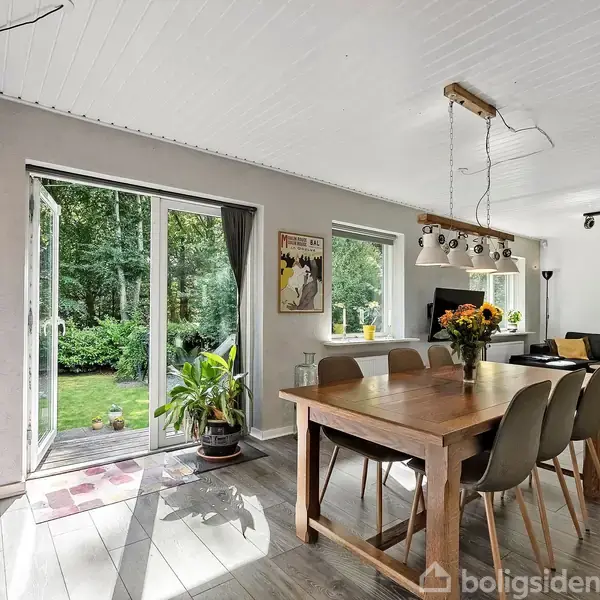 Træbord med seks stole, blomsterbuketter og lamper ovenover. Franske døre åbne til have med græs og buske. Stue med sofa og lyse træmøbler.