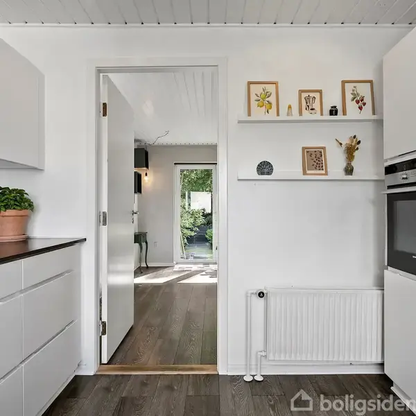 Et køkken med hvide skabe og en indbygget ovn åbner mod en lys stue. Grøn plante og billeder på hylderne tilføjer dekoration. Der ses trægulve og en radiator.