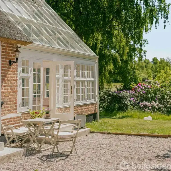 En solrig terrasse med et bord og to stole foran et hus med glasveranda. Omkringliggende have med græs, buske og træer i baggrunden.