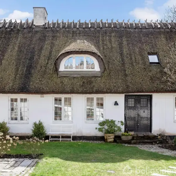 Hus med stråtag, hvide vægge og sorte døre står i en have med græsplæne og blomsterkrukker, omgivet af åbne hvide dobbeltdøre der giver udsyn til en blå himmel.