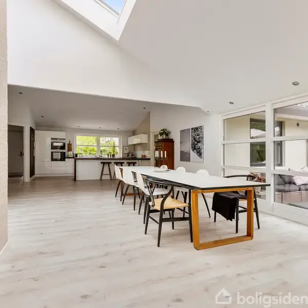 Spisebord med stole står i en rummelig, moderne stue med lyse gulve. Pejsen er tændt til venstre, og store vinduer giver udsigt til haven. En plante i kurv står i hjørnet.