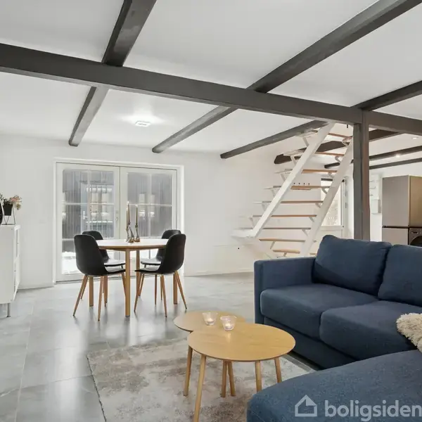 En moderne stue med en blå sofa i forgrunden, runde borde, og en trappe der fører til en øvre etage. Baggrunden har et spisebord foran store vinduer.