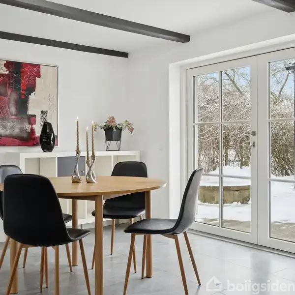 Rundt træbord med fire sorte stole står i et lyst rum. På bordet er der høje lysestager. En moderne kunstplakat hænger på væggen. Udenfor ses snedækket terræn.