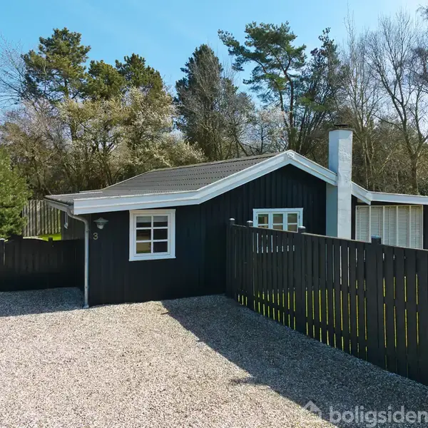 Et sortmalet træhus står stille i en skovomgivet baggrund. En grusbelagt indkørsel leder op til huset, som er omgivet af et hegn. Tallene "8" kan ses ved døren.