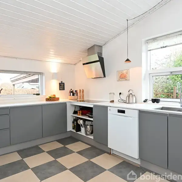 Køkken med grå skabe og skuffer, hvid bordplade, underbelysning, store vinduer med udsigt til have, og sort-hvid ternet gulv. Kaffemaskine og brødrister står ved vinduet.