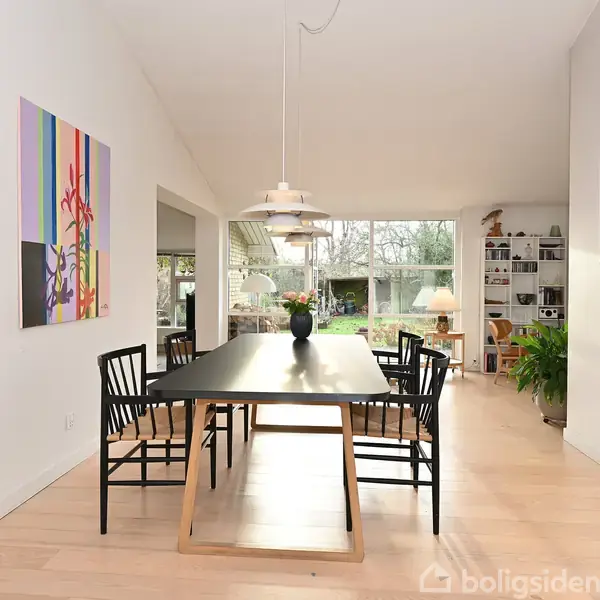 Et sort spisebord med seks sorte stole står centralt i et lyst rum. Over bordet hænger en lampe, og der er et stort vindue mod haven. Malerier pryder væggene.