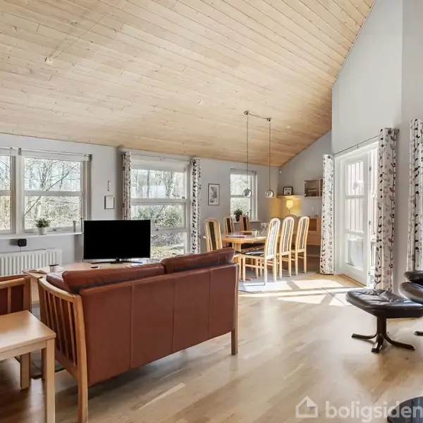 Stue med lyse trægulve og loft, hvor en brun sofa og lænestol er central. Et spisebord med stole står ved siden af vinduer med udsigt til en skov.