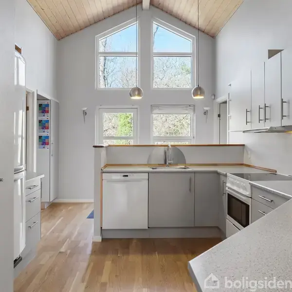 Køkken med lyse trægulve under loftsloft, hvor to hængelamper lyser rummet. Store vinduer bag vasken giver udsigt til træer. Dekorationer hænger på væggene, en med teksten "Coffee".