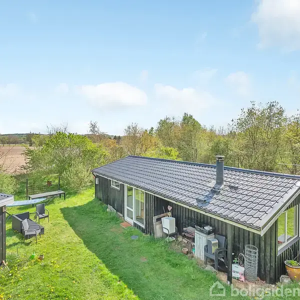 Et sort træhus med gråt tag står stille i et grønt haveområde omgivet af træer og buske. En lille terrasse til venstre har havemøbler.