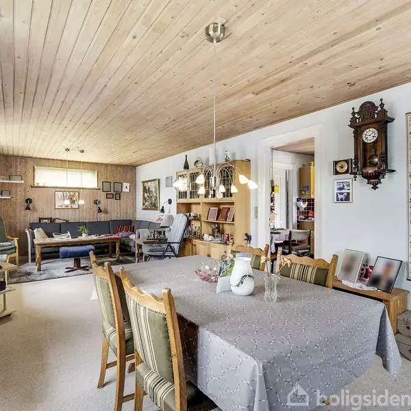 Spisestue med lang, dækket bord og stole, dekorativ væg med ur og kunst. I forlængelse er en opholdsstue med sofaer og reoler, i et rum med træloft og lyse vægge.
