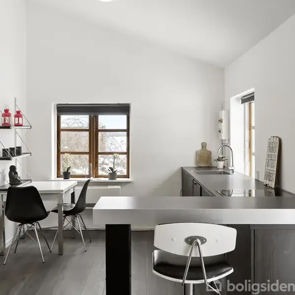 Et moderne køkken med mørke trægulve og hvide vægge, der rummer en sort køkkenø og barstol, sort spisebord med stole, og hylder med dekorative genstande langs væggen.