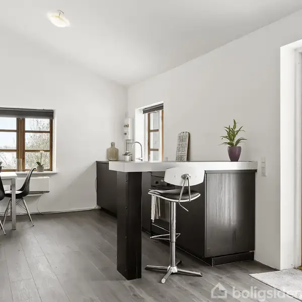 Køkken med mørke trægulve; høj barstol ved køkkenø; sorte skabe; to vinduer med udsigt til snebeklædte buske; åben dør til terrasse; på væggen hænger sorte hylder og et lille arbejdsbord.