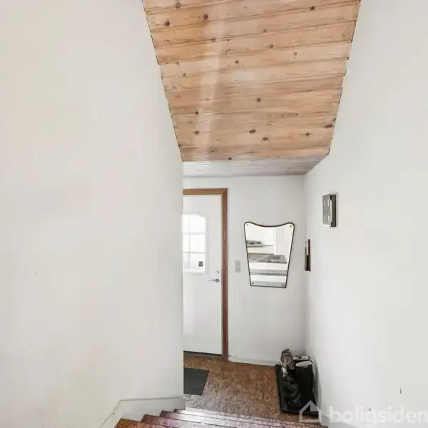 Trappe op ad en gang, fører til en hvid dør med vindue i en lys entre. En spejl hænger på væggen, ved siden af en lille opslagstavle.